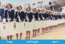 ცა, როგორც მუზა – რატომ გვხიბლავს ფრენა ასე ძალიან? A row of vintage flight attendants in elegant blue and white uniforms holding parasols in front of a classic propeller airplane.