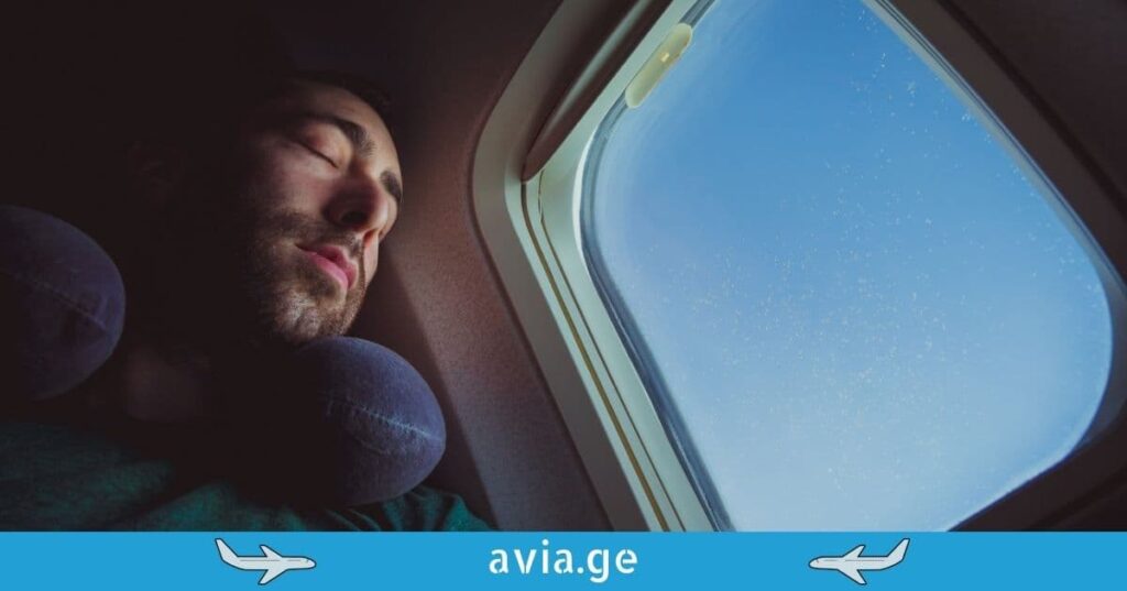 A man with a travel neck pillow sleeping next to an airplane window