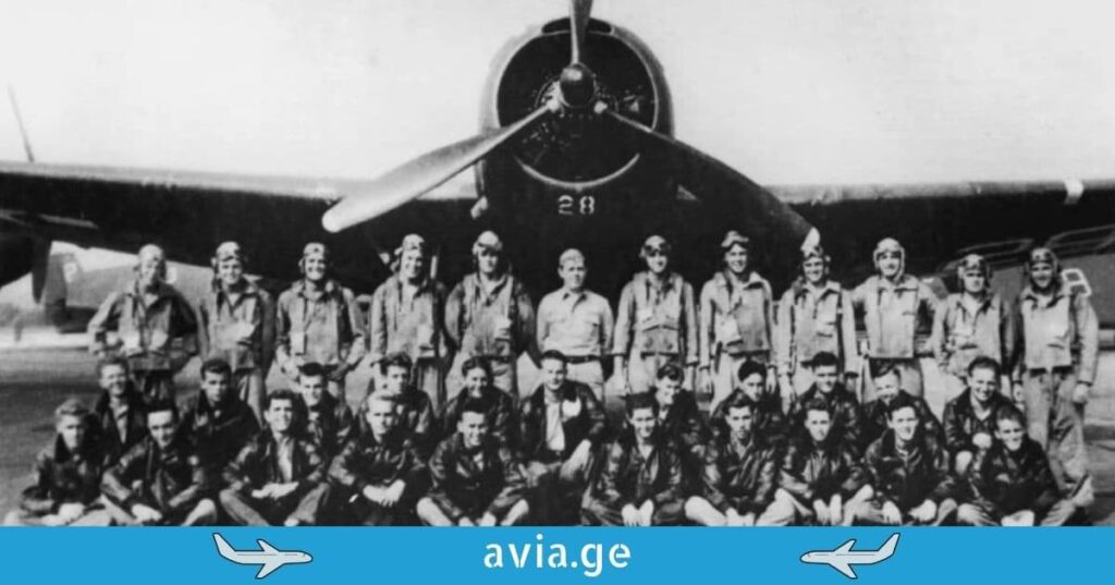 A vintage black and white photograph shows a large group of aviators posing in front of a propeller aircraft.