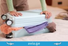 Wizz Air-ის ბარგის პოლიტიკა: ყველაფერი, რაც მგზავრობამდე უნდა შეამოწმოთ A person packing a light blue hardshell suitcase on a bed, struggling to close it over colorful clothes, featuring the avia.ge logo footer.