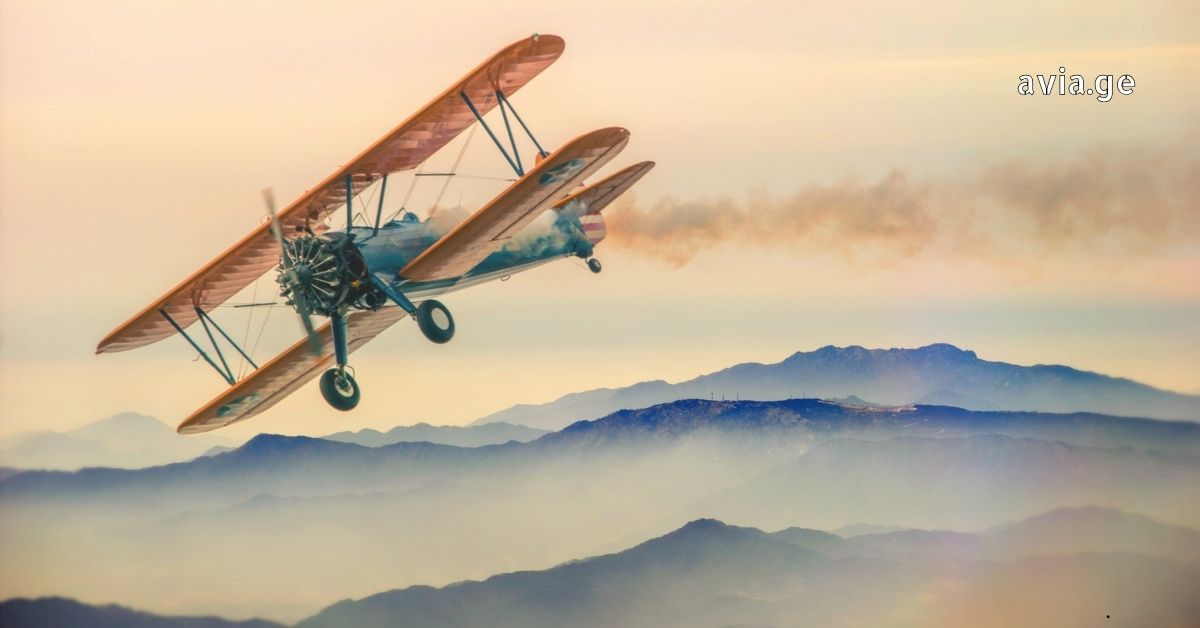 A vintage biplane flying over misty mountains during sunset.