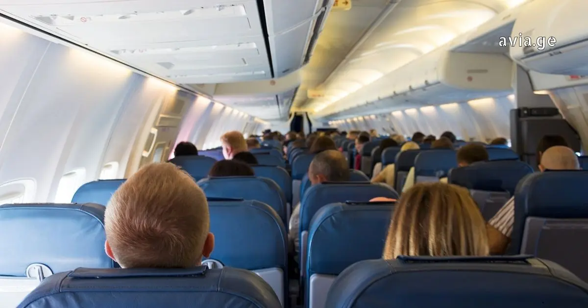Interior view of a crowded commercial flight cabin with passengers seated in blue chairs.