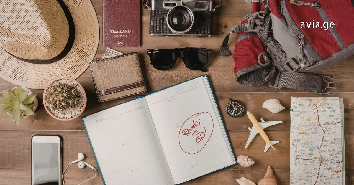 Flat lay of travel essentials including passport, camera, notebook with 'Ready to Go!' text, and miniature airplane on a wooden table.