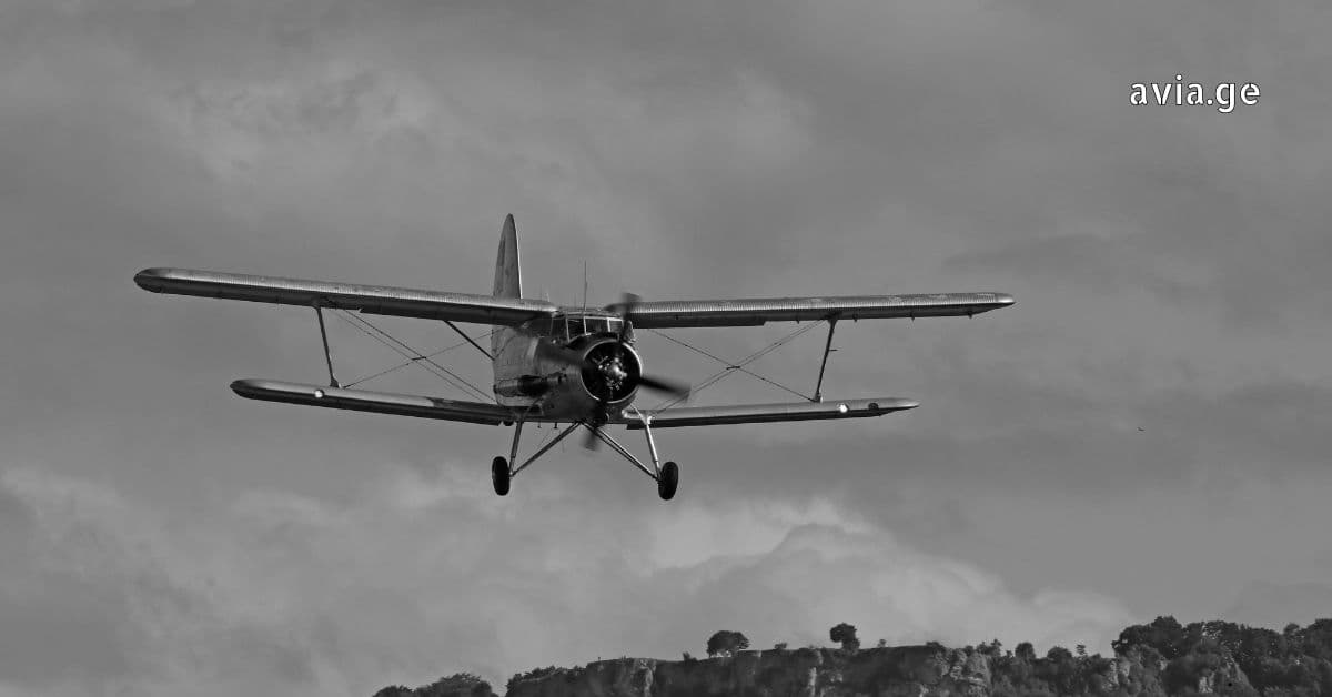 A black and white photo of a vintage propeller biplane in flight against a cloudy sky, seen from a front-facing angle.
