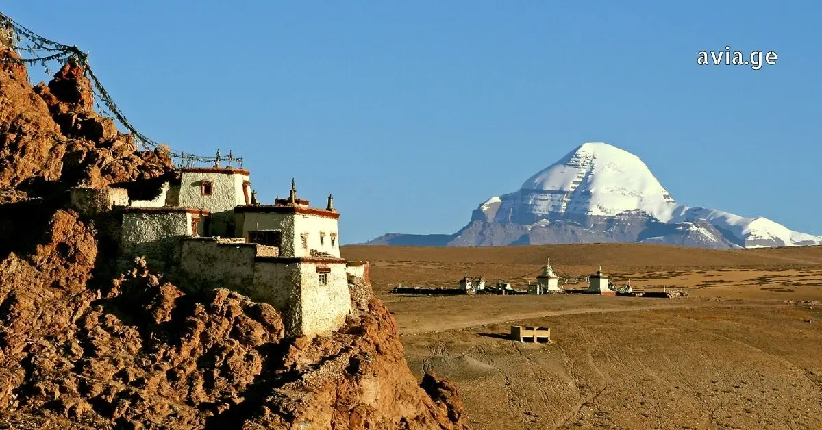 Traditional white Tibetan stone buildings perched on a rocky cliffside, overlooking a vast plain with the snow-covered Mount Kailash peak in the distance under a clear blue sky.