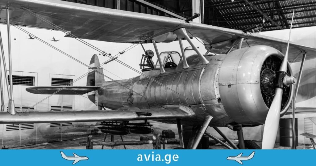 A black and white photo of a classic vintage biplane with a large front propeller, parked inside an aviation museum or hangar.