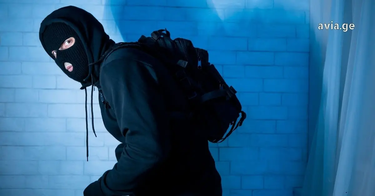 მამაკაცი შავ ნიღაბსა და ჰუდში, ზურგჩანთით, იყურება უკან ლურჯად განათებული აგურის კედლის ფონზე, "avia.ge" ლოგოთი.