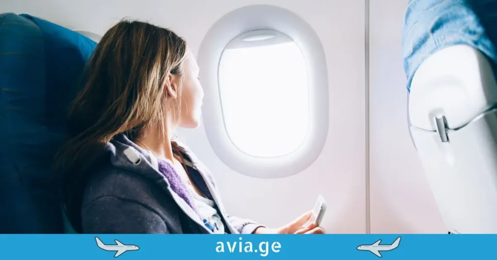 გოგონა თვითმფრინავში ილუმინატორთან, რომელიც ავიაბილეთების გარეშე მოგზაურობაზე ფიქრობს.