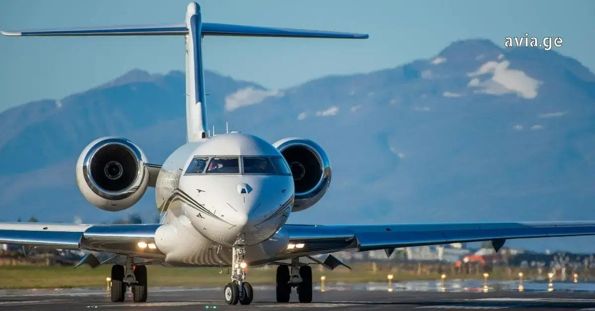 თეთრი კერძო თვითმფრინავი ასაფრენ ბილიკზე, მთების ფონზე, ავია.ჯი-ს ლოგოთი.